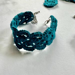 May include: A teal blue crocheted bracelet with a silver clasp. The bracelet is made of a delicate, openwork design.