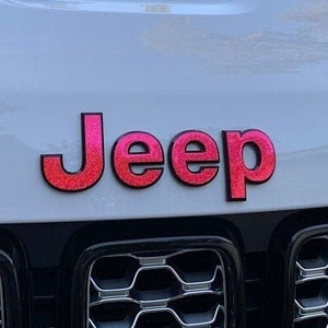 Könnte beinhalten: Ein weißer Jeep mit einem pinkfarbenen Glitzer-Jeep-Logo auf der Front. Der Jeep hat einen schwarzen Kühlergrill mit einem Wabenmuster.