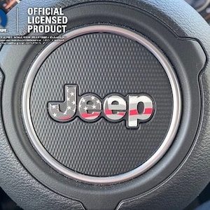 May include: A black steering wheel with a silver trim ring and a red, white, and blue American flag design on the Jeep logo. The text "OFFICIAL LICENSED PRODUCT" is printed above the logo.