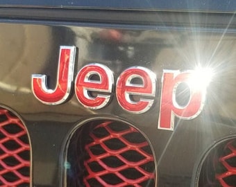 May include: A black Jeep grille with a red mesh insert and a chrome Jeep logo with red lettering. The logo reads "Jeep".