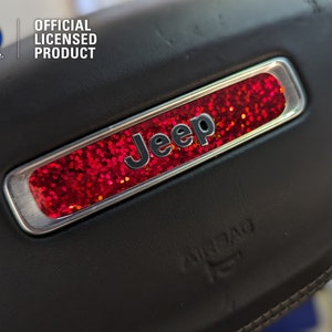 May include: A close-up of a black leather car seat with a silver trim and a red glitter "Jeep" logo. The logo is surrounded by a silver trim and has the words "Official Licensed Product" above it.