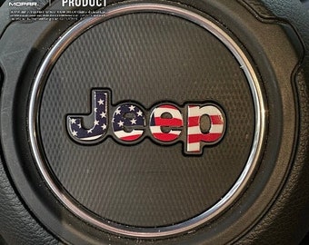 May include: A black Jeep steering wheel cover with a silver trim ring. The Jeep logo is in the centre of the cover and is decorated with an American flag design.