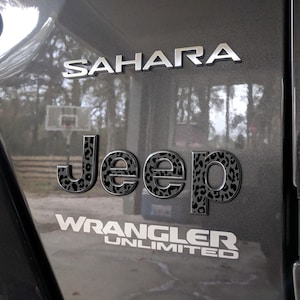 May include: Close-up of a gray vehicle panel with chrome lettering that reads "SAHARA" and "WRANGLER UNLIMITED." The word "JEEP" is in a bold font with a black and gray leopard print pattern. The background shows a basketball hoop.