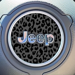 May include: Close-up of a Jeep steering wheel emblem. The emblem features the word "Jeep" in a chrome-like font with a leopard print background. The emblem is set in a circular frame, surrounded by a gray steering wheel.