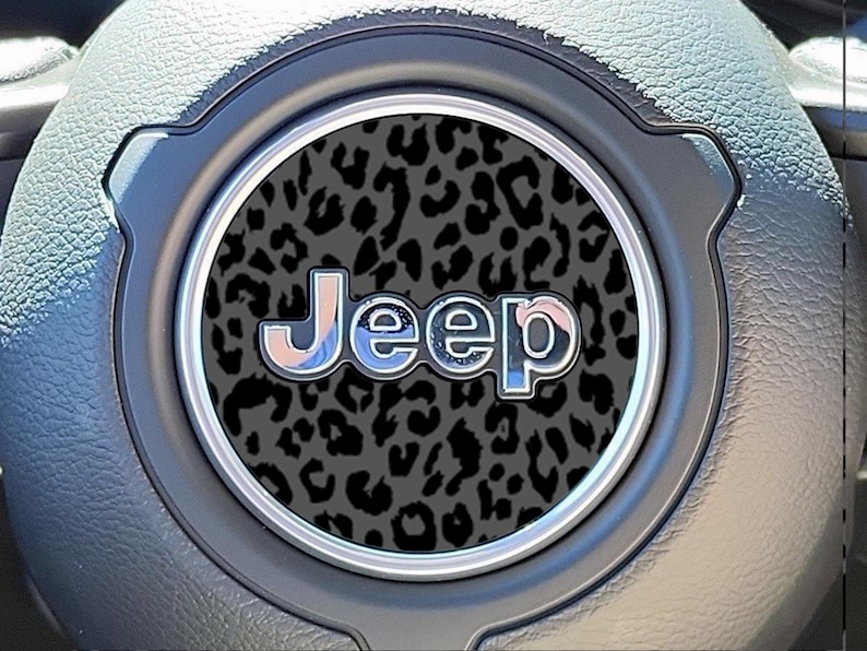 May include: Close-up of a Jeep steering wheel emblem. The emblem features the word "Jeep" in a chrome-like font with a leopard print background. The emblem is set in a circular frame, surrounded by a gray steering wheel.