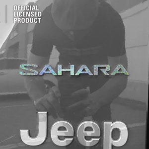 Pu&ograve; includere: Una Jeep nera con un emblema "Jeep" argentato e un emblema "Sahara" argentato con finitura olografica. L'emblema "Sahara" &egrave; sopra l'emblema "Jeep". La Jeep &egrave; parcheggiata in un garage.