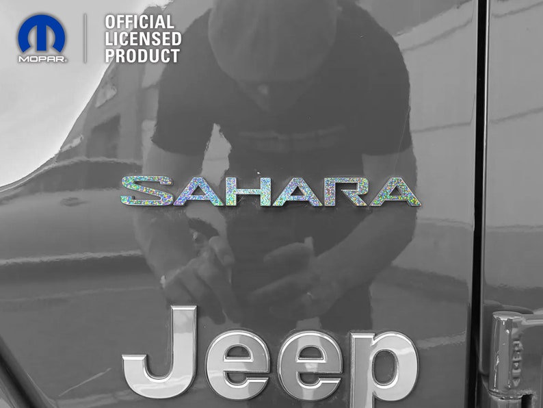 Pu&ograve; includere: Una Jeep nera con un emblema "Jeep" argentato e un emblema "Sahara" argentato con finitura olografica. L'emblema "Sahara" &egrave; sopra l'emblema "Jeep". La Jeep &egrave; parcheggiata in un garage.