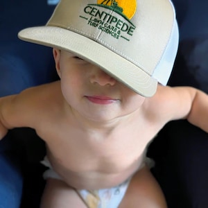 May include: A young child wearing a beige and white baseball cap with the text "CENTIPEDE - LAWN CARE & TURF SCIENCES" embroidered on the front. The child is sitting on a dark blue surface.