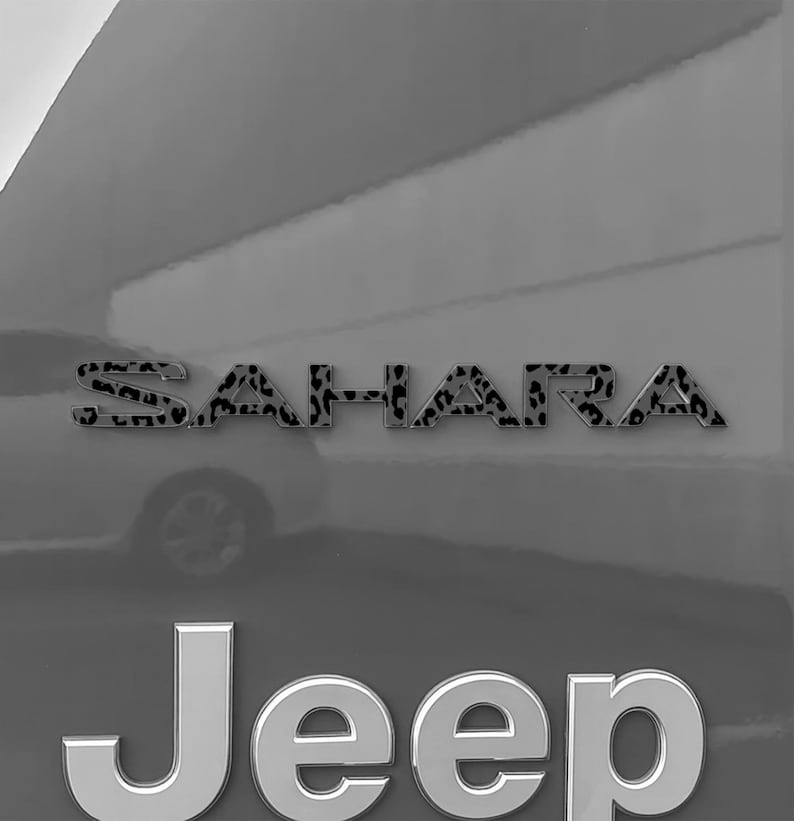 May include: Close-up of a gray vehicle with the word "Sahara" in leopard print lettering above the word "Jeep" in silver block letters. The vehicle's body is a solid gray color.