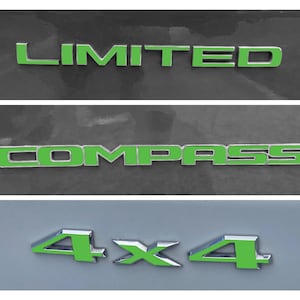 Peut inclure: Gros plan sur trois emblèmes automobiles. Le premier lit "LIMITED" en lettres vert vif. En dessous, le mot "COMPASS" est affiché dans le même vert. En bas, l'emblème "4X4" est également en vert.