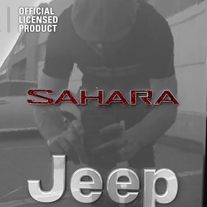 Pu&ograve; includere: Un emblema "SAHARA" rosso glitterato su una Jeep grigia con un emblema "Jeep" argentato sotto. L'immagine &egrave; un primo piano della parte posteriore del veicolo.