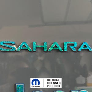 Pu&ograve; includere: Un emblema "SAHARA" blu turchese e argento con un logo Mopar argento e il testo "PRODOTTO LICENZIATO UFFICIALE" sotto.