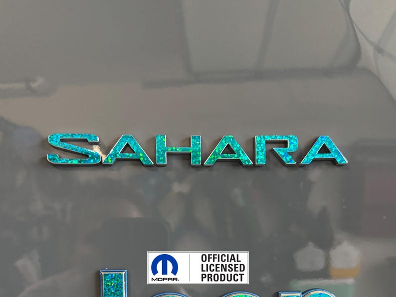 Pu&ograve; includere: Un emblema "SAHARA" blu turchese e argento con un logo Mopar argento e il testo "PRODOTTO LICENZIATO UFFICIALE" sotto.
