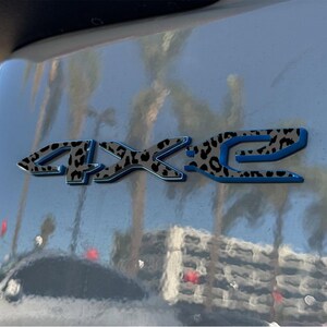 May include: Close-up of a vehicle's exterior with a 4X2 emblem. The emblem features a leopard print design with a blue outline. The background shows reflections of palm trees and other vehicles.