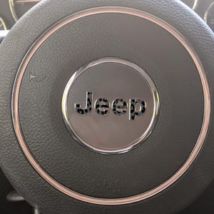 May include: Close-up of a black leather steering wheel with a silver center. The center features the word "Jeep" in a stylized font. A silver ring surrounds the center, and the horn symbol is visible on the left side.
