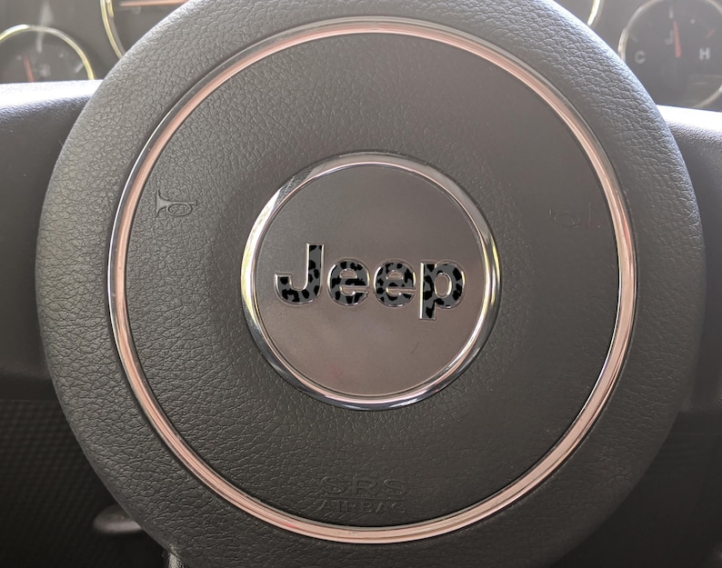 May include: Close-up of a black leather steering wheel with a silver center. The center features the word "Jeep" in a stylized font. A silver ring surrounds the center, and the horn symbol is visible on the left side.