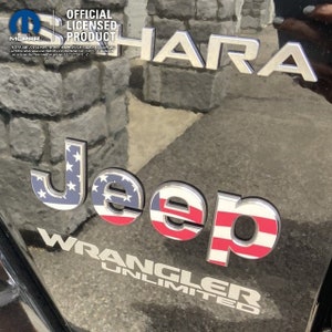 May include: A silver Jeep Wrangler Unlimited emblem with a red, white, and blue American flag design. The emblem is attached to a black vehicle. The text "Jeep" is in a bold, white font with the American flag design behind the letters. The text "Wrangler Unlimited" is in a smaller, white font below the Jeep emblem.