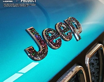 May include: A close-up of a turquoise Jeep with a black and silver "Jeep" logo with iridescent glitter. The logo is on the front of the car, above the grille.