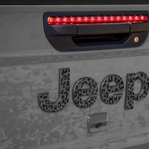 May include: Close-up of a gray vehicle's tailgate with a black handle and a red LED light strip. The word "Jeep" is displayed in chrome letters with a leopard print pattern. A small camera is visible below the letters.