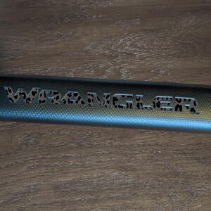 May include: A close-up of a blue metal bar with the word "WRANGLER" etched into it. The letters are filled with a leopard print pattern. The bar has a textured surface and is set against a wooden background.