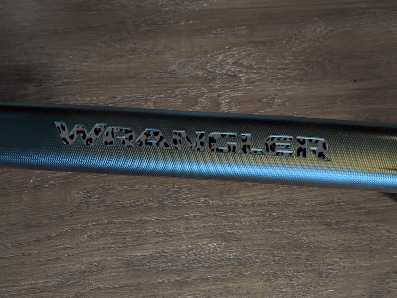 May include: A close-up of a blue metal bar with the word "WRANGLER" etched into it. The letters are filled with a leopard print pattern. The bar has a textured surface and is set against a wooden background.