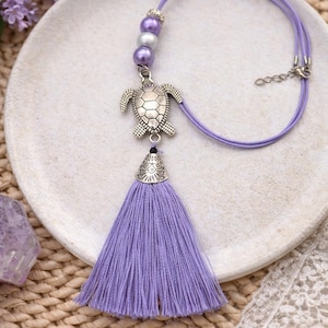 Peut inclure: Un collier lavande avec un pendentif tortue argenté et un pompon assorti. Le collier est composé d'un cordon avec des perles et d'un capuchon en métal décoratif sur le pompon. Le bijou est présenté sur une assiette blanche.