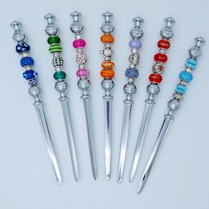 Może przedstawiać: Siedem otwieraczy do listów ze srebrnymi uchwytami i ozdobnymi, koralikowymi wierzchami. Koraliki są w różnych kolorach, w tym niebieskim, zielonym, różowym, pomarańczowym, fioletowym i czerwonym. Otwieracze do listów są ułożone na białej powierzchni.