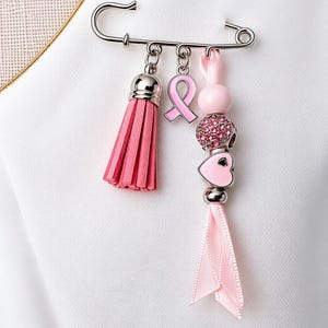 Pode incluir: Um broche de alfinete prateado com três pingentes pendurados: uma borla de camurça rosa, um laço de conscientização de esmalte rosa e um fio com contas rosas, uma conta com strass, um coração rosa e uma fita rosa claro.