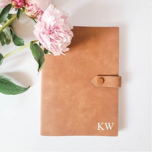 Puede incluir: Un diario de cuero marrón claro con cierre de botón y las iniciales "KW" en blanco. El diario es rectangular y tiene una superficie texturizada. Peonías rosas y hojas verdes en la esquina superior izquierda.
