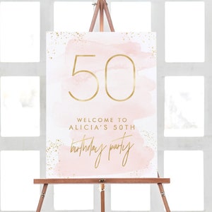 May include: A sign on a wooden easel with the number 50 in gold script. The sign has a pink watercolor background with gold flecks and text that reads "Welcome to Alicia's 50th Birthday Party."