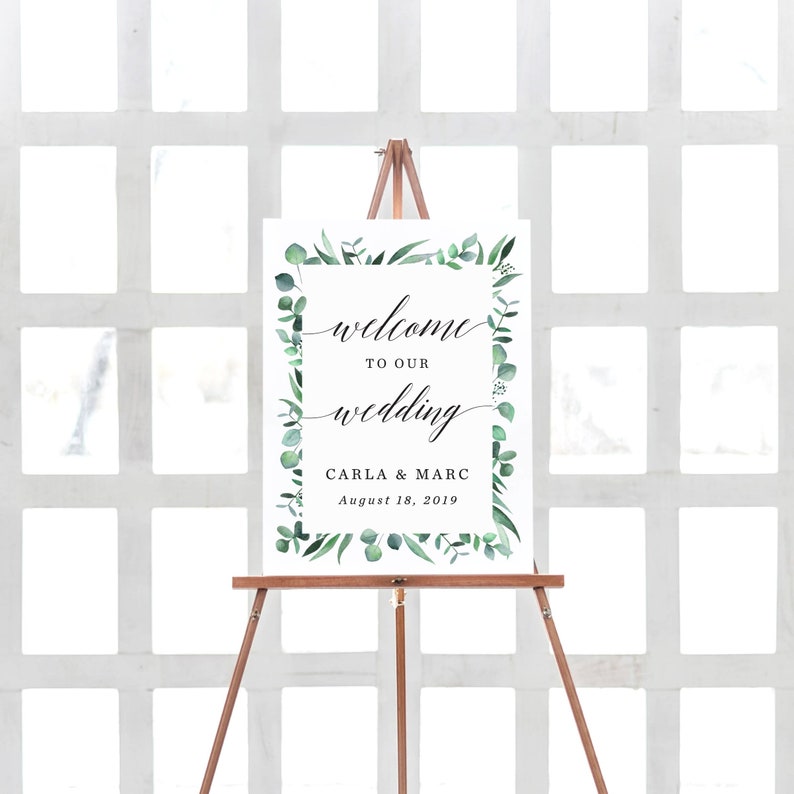 May include: A wedding welcome sign on a wooden easel. The sign has a white background with the words "welcome to our wedding" in elegant script. The sign is framed with green leaves and the names Carla & Marc and the date August 18, 2019.