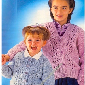 May include: Two young girls wearing knitted cardigans. One girl is wearing a light blue cardigan with a white collared shirt and a plaid skirt. The other girl is wearing a pink cardigan over a striped shirt and navy blue pants.