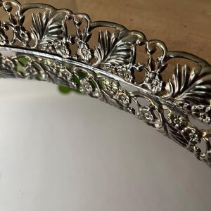 Op de afbeelding: Close-up van een zilverkleurige decoratieve schaal met een sierlijk bloemen- en bladerontwerp rond de rand. De rand van de schaal is ingewikkeld gedetailleerd en toont een vintage esthetiek. De schaal wordt waarschijnlijk gebruikt om te serveren of tentoon te stellen.