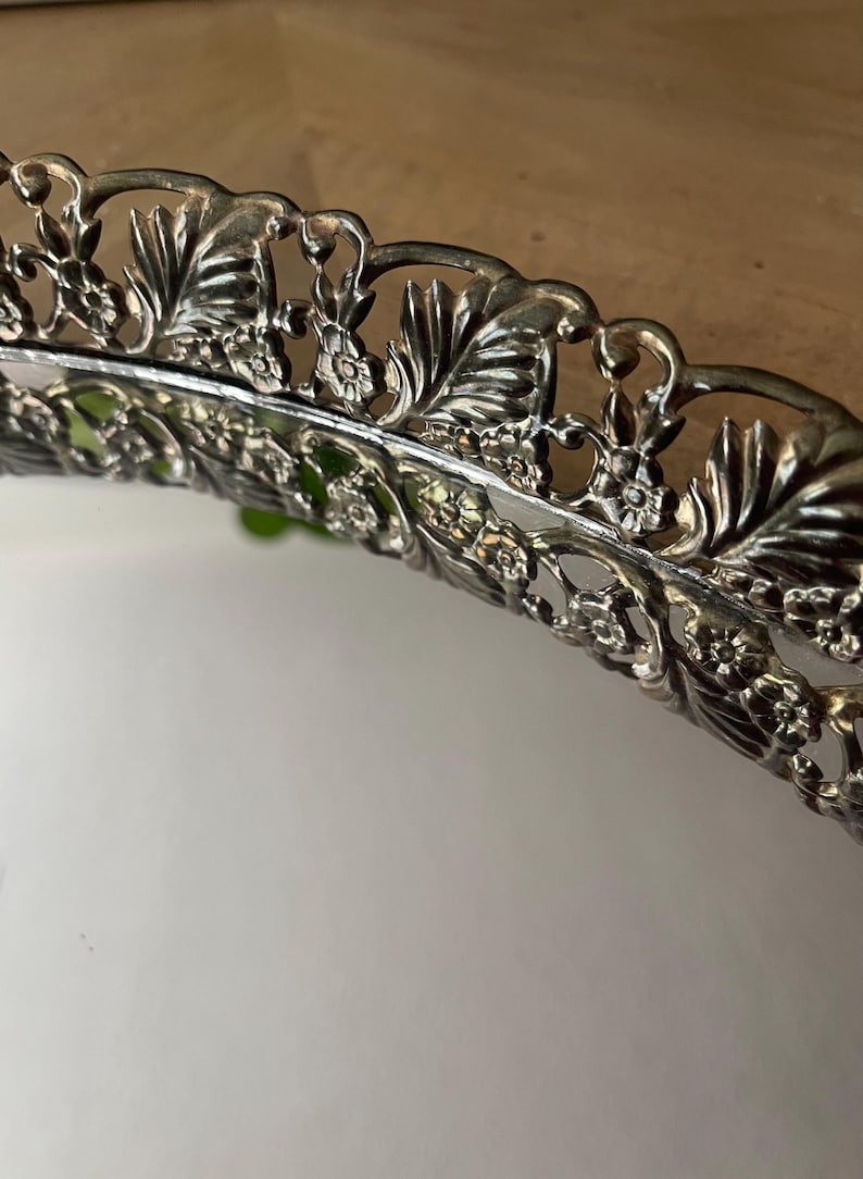 Op de afbeelding: Close-up van een zilverkleurige decoratieve schaal met een sierlijk bloemen- en bladerontwerp rond de rand. De rand van de schaal is ingewikkeld gedetailleerd en toont een vintage esthetiek. De schaal wordt waarschijnlijk gebruikt om te serveren of tentoon te stellen.