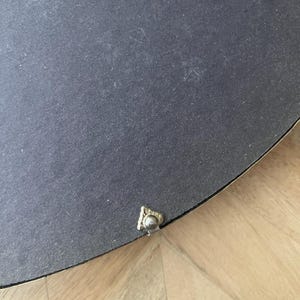 Op de afbeelding: Een close-up van de achterkant van een rond, zwart object. Een klein, sierlijk, goudkleurig metalen stukje is aan de rand bevestigd. Het object rust op een lichtgekleurd houten oppervlak.