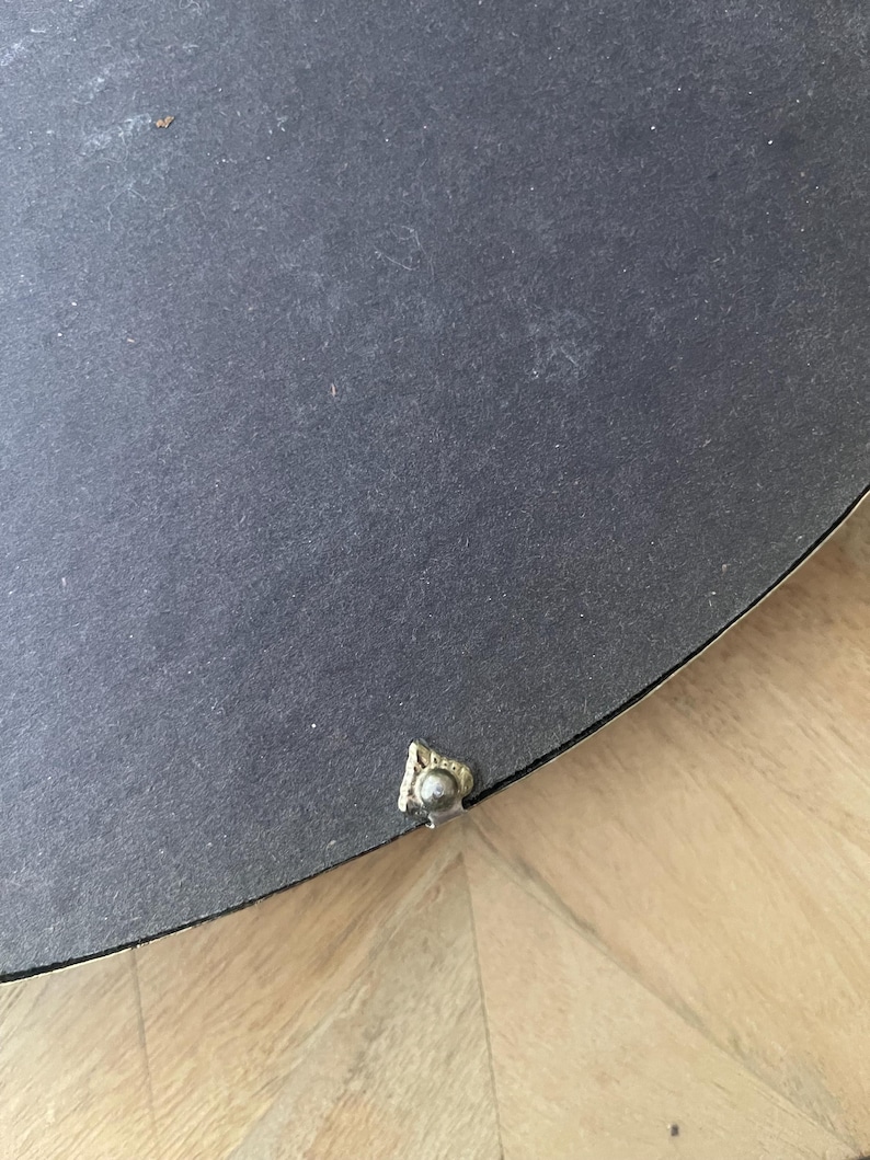 Op de afbeelding: Een close-up van de achterkant van een rond, zwart object. Een klein, sierlijk, goudkleurig metalen stukje is aan de rand bevestigd. Het object rust op een lichtgekleurd houten oppervlak.