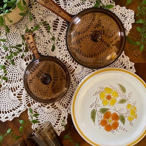May include: Two amber-colored glass saucepans with matching lids, two matching amber glass mugs, and a round white ceramic plate featuring a yellow and orange floral pattern with a yellow border, arranged together on a white crocheted lace doily.