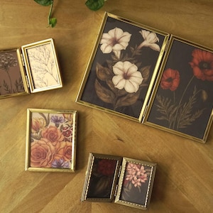 May include: A collection of small, gold-framed pictures with floral artwork. The images feature various flowers, including white petunias, red poppies, and roses, set against a dark background. The frames are rectangular and have a decorative edge.