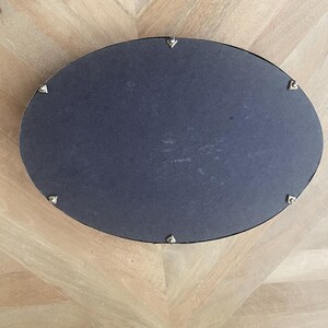 Op de afbeelding: Een ovale spiegel met een donkergrijze oppervlakte en een zilverkleurig frame. Het frame is versierd met kleine, decoratieve zilveren accenten. De spiegel staat op een lichtgekleurd houten oppervlak.