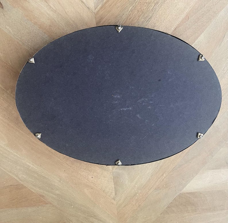 Op de afbeelding: Een ovale spiegel met een donkergrijze oppervlakte en een zilverkleurig frame. Het frame is versierd met kleine, decoratieve zilveren accenten. De spiegel staat op een lichtgekleurd houten oppervlak.