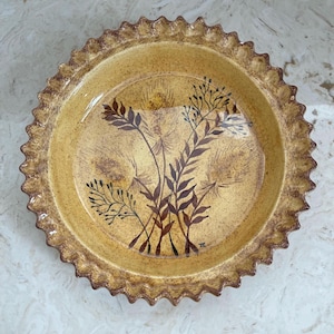 May include: A round, ceramic pie plate with a scalloped edge. The plate is a warm yellow color with a brown floral design in the center. The design features stylized leaves and stems. The plate has a rustic, handmade appearance.