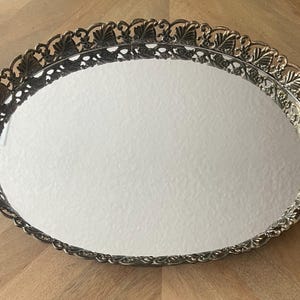 Op de afbeelding: Een ovale, zilverkleurige ijdelheidsschaal met een spiegeloppervlak. De schaal heeft een decoratieve, opengewerkte rand met een herhalend bladerenontwerp. De schaal is leeg en ligt op een houten oppervlak.