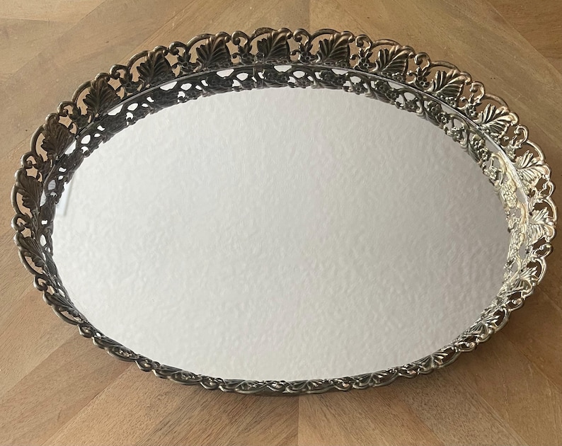 Op de afbeelding: Een ovale, zilverkleurige ijdelheidsschaal met een spiegeloppervlak. De schaal heeft een decoratieve, opengewerkte rand met een herhalend bladerenontwerp. De schaal is leeg en ligt op een houten oppervlak.