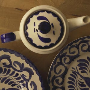 May include: Overhead view of a white ceramic teapot and two matching plates. The teapot has a blue handle and spout, with a blue and white patterned lid. The plates feature a similar blue and white floral design, perfect for serving tea or other beverages.