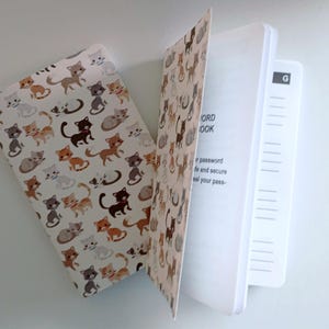 May include: A cat-themed password book with a white cover featuring various cartoon cat illustrations in brown, gray, and orange. The open book reveals lined pages and a tabbed section for easy organization. The text "PASSWORD BOOK" is visible.