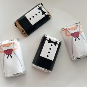 May include: Four small chocolate bars wrapped in silver foil with printed images of a bride and groom. The bride is wearing a white dress and holding a red bouquet. The groom is wearing a black tuxedo with a white shirt and a black bow tie.