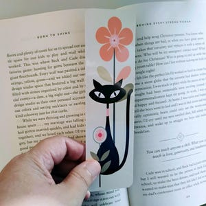 May include: A rectangular bookmark featuring a black cat illustration with white eyes and a pink collar. Above the cat is a large coral-colored flower with green leaves, and a smaller pink circular flower sits to the left of the cat's body.