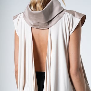 Puede incluir: Una blusa blanca sin mangas con cuello de pico y una gran espalda abierta. La blusa está hecha de una tela suave y drapeada.