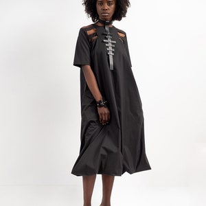 May include: A black midi dress with a cut-out design on the chest and a black leather harness with silver buckles. The dress has a loose fit and a flowing skirt. The model is wearing black platform sandals.