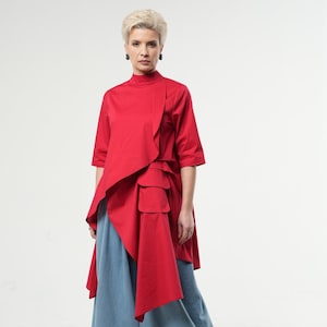 Pode incluir: Um top vermelho assimétrico com gola alta e um grande bolso na frente. O top é combinado com calças largas azul claro.